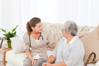 A home health provider talks to an elderly woman sitting on her couch.
